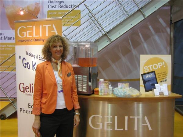 A woman with curly hair in an orange jacket stands beside a Gelita banner and a Gelita counter holding a beverage dispenser, product samples and a sign about winning an iPad.