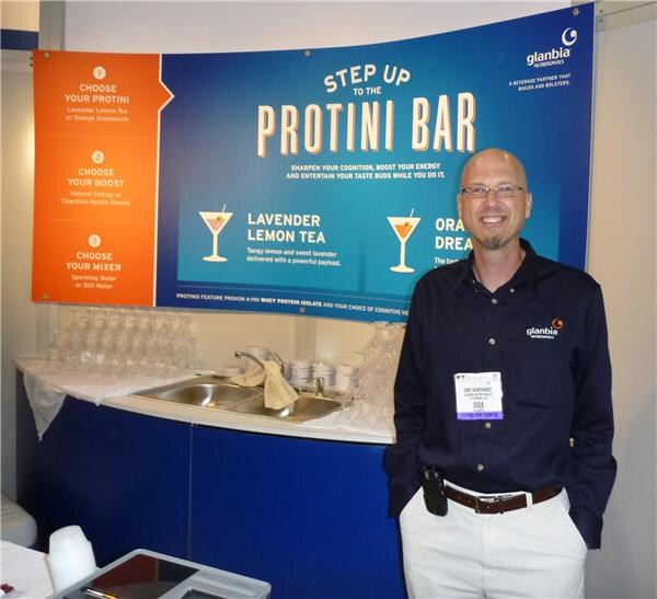 A smiling bald man with glasses and a goatee stands in a Glanbia Nutritionals shirt in front of the Step Up to the Protini Bar sign, with a small sink and glassware behind him.