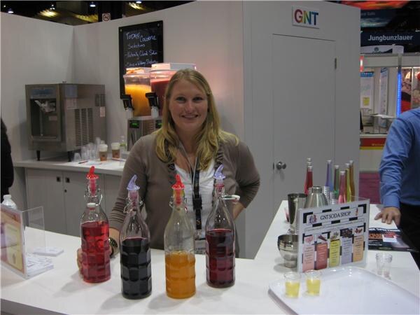 A smiling woman with blonde hair stands behind a counter holding four bottles of red, dark and orange syrups, with a GNT Soda Shop sign and a GNT logo behind her.