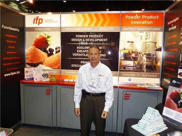 A man in a white shirt stands in the IFP booth in front of panels reading Powder Product Innovation, with images of strawberries, orange powder and a processing plant.