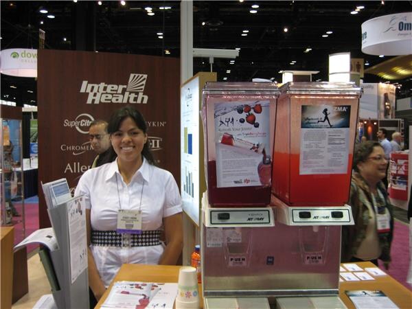 A smiling woman in a white blouse stands at the InterHealth booth beside two beverage dispensers filled with red drinks, with the InterHealth logo on the wall behind her.