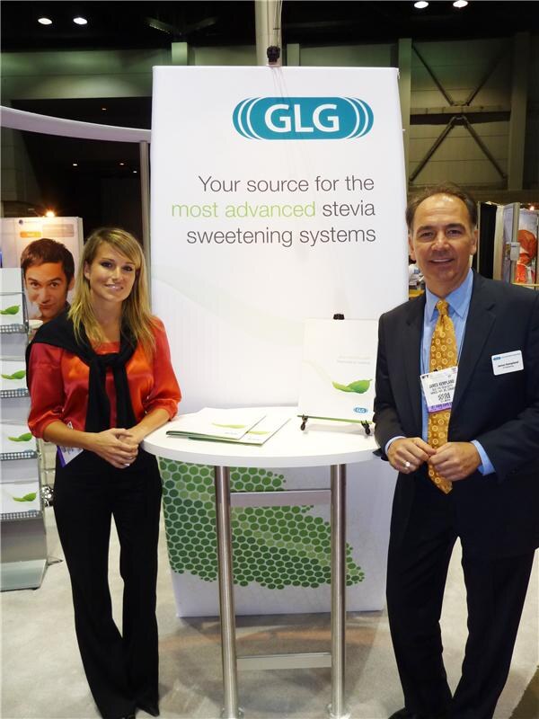 James Kempland, in a dark suit and yellow tie, stands at right beside a woman in an orange top at a small table in front of a GLG banner about stevia sweetening systems.
