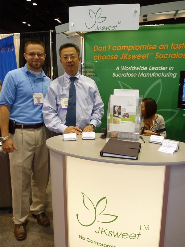 Two men with badges stand at a round JKsweet counter in front of a green wall reading Don't compromise on taste, choose JKsweet Sucralose, while a woman sits behind the counter.