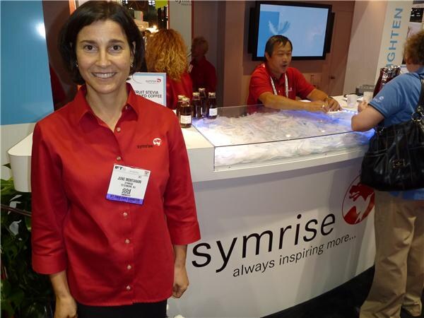June Montanari, in a red Symrise shirt and event badge, smiles at left in front of the Symrise booth. A man in red stands behind the counter, with bottles and ice on display.