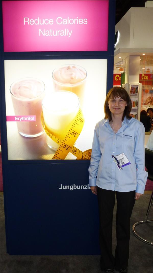 A woman in a light blue Jungbunzlauer shirt stands beside a lit display reading Reduce Calories Naturally, which shows glasses of smoothie wrapped with a tape measure and an Erythritol label.