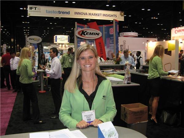 A smiling woman with long blonde hair in a green jacket stands at the Kemin booth, with the Kemin sign overhead and other exhibitors and a Watson sign in the background.