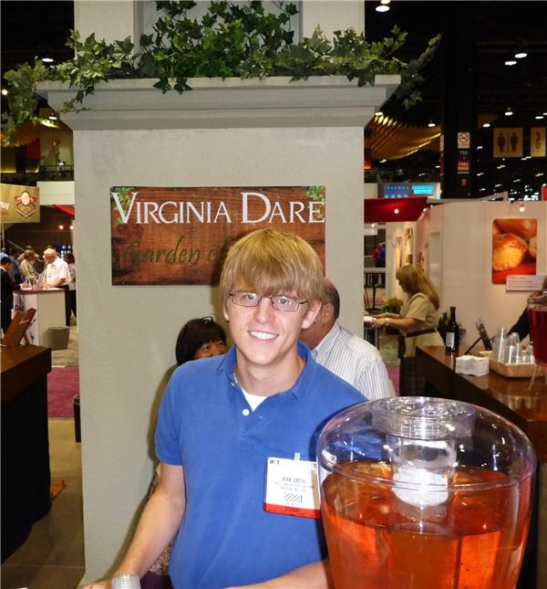 Kim Smith, a smiling young man with glasses and a blue polo shirt, stands beside a Virginia Dare sign and a large glass dispenser of orange-red drink, with the show floor behind him.