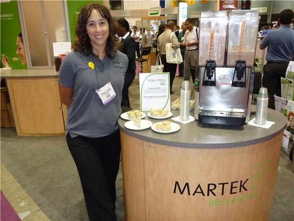 A smiling woman in a gray polo stands next to a round Martek counter holding a beverage dispenser, a Today's Menu sign and plates of small baked items, with attendees behind her.