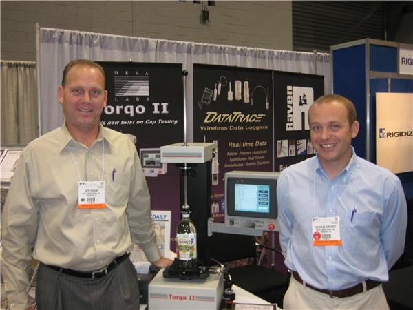 Two smiling men with badges stand in the Mesa Labs booth beside a Torqo II cap testing instrument holding a bottle, with DataTrace and Raven signs behind them.