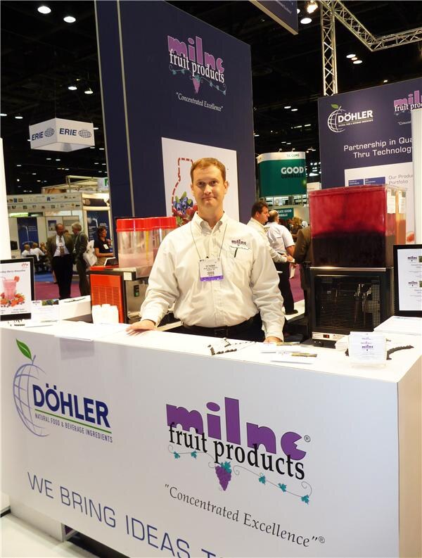 A man in a white shirt and badge stands behind a counter with Döhler and Milne Fruit Products signs, with beverage dispensers on either side.