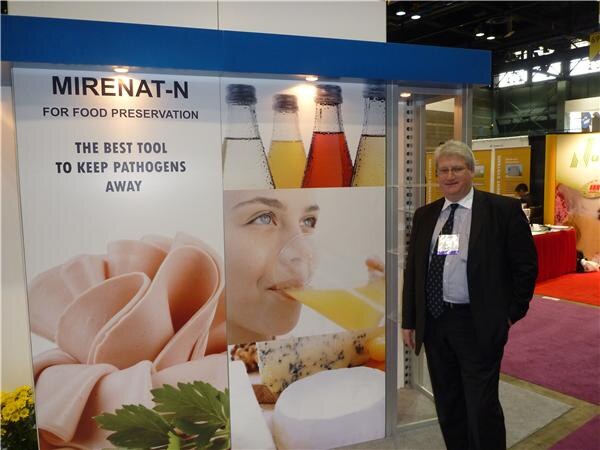 A man in a dark suit with a name badge stands beside a booth wall reading 'Mirenat-N for food preservation', with images of bottled juices, sliced deli meat, cheese and a woman drinking juice.