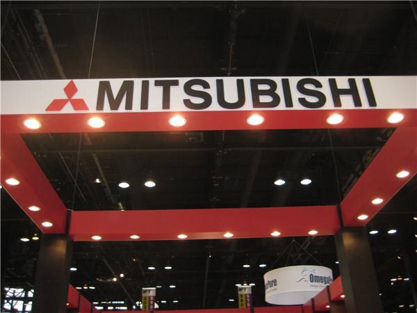 Overhead red booth frame with a white Mitsubishi sign and red three-diamond logo, lit by recessed ceiling lights, with other exhibit signs visible in the background.