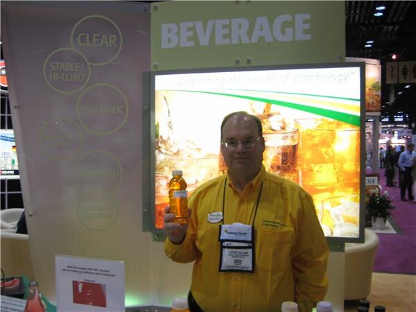 A man in a yellow shirt and glasses holds up a bottle of amber beverage in front of a lit Beverage display at the National Starch booth.