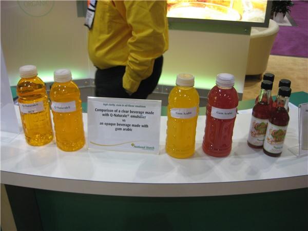Counter at the National Starch booth with bottles of yellow, orange and red beverages beside a sign comparing a clear Q-Naturale drink with an opaque gum arabic drink.