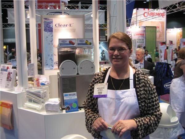 Nora Watson, a woman with glasses wearing a white apron over a patterned jacket, stands in front of a Clear-E display with a Taylor frozen beverage machine. A Caremoli banner hangs behind her.
