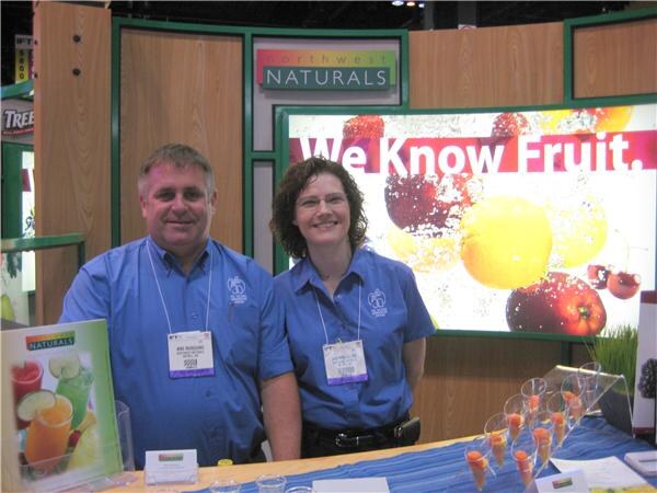 A man and a woman in blue shirts and badges stand at the Northwest Naturals booth, with a We Know Fruit sign behind them and small sample cups on the table.