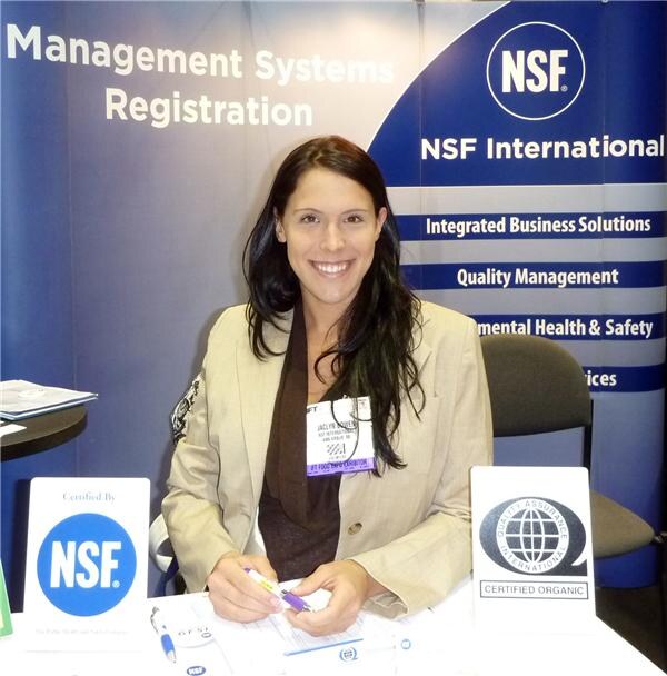 A smiling woman with long dark hair sits at the NSF International booth holding a pen, with NSF and Quality Assurance International Certified Organic signs on the table.