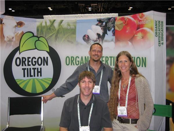 Two men and a woman wearing badges pose in front of an Oregon Tilth banner about organic certification and education, with images of a cow, fields and apples.