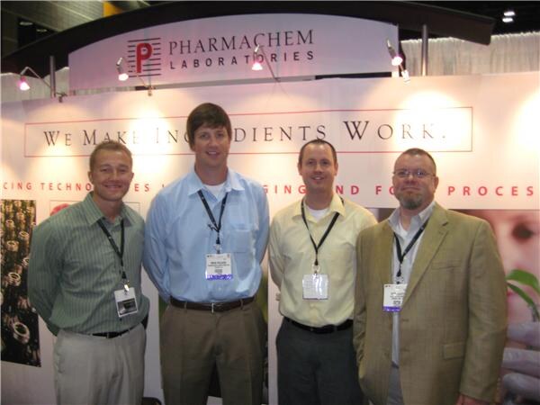 Four men wearing badges stand in front of the Pharmachem Laboratories booth, under a sign reading We Make Ingredients Work.