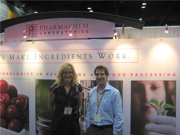 A blonde woman with glasses and a man in a light blue shirt stand in front of the Pharmachem Laboratories booth and its We Make Ingredients Work sign.