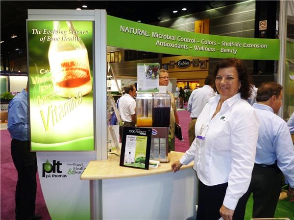 A smiling woman in a white shirt stands beside the P.L. Thomas booth, which has a green banner and a lit panel about bone health, with other attendees behind her.