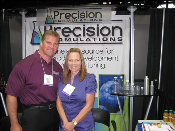 A man in a maroon polo and a woman in a purple blouse stand in front of a Precision Formulations banner, with bottles on a small table beside them.