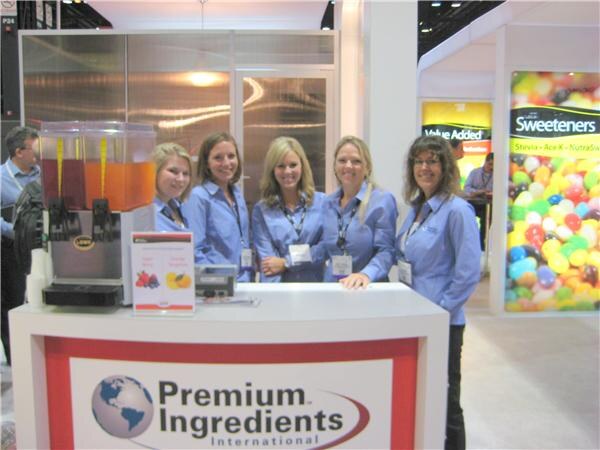 Five smiling women in blue button-up shirts and lanyards stand behind a booth counter with a beverage dispenser. The counter displays the Premium Ingredients International name and globe logo.