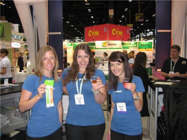 Three women in blue T-shirts with badges hold small cups of drink and a green and white packet. Behind them are REIPU and China signs, and a man stands at a counter on the right.