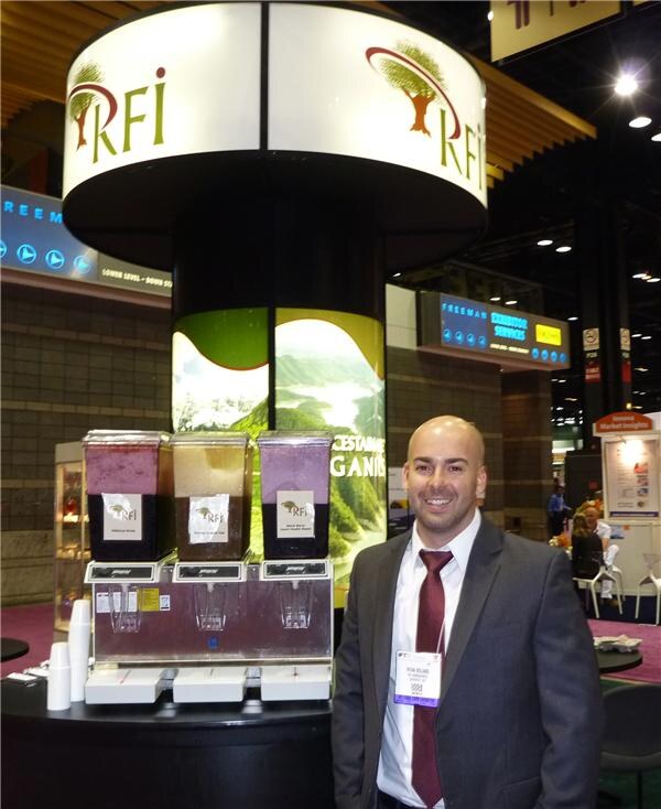 A bald man in a gray suit, maroon tie and badge stands smiling beside a booth with three beverage dispensers. A round RFI sign with a tree logo hangs overhead.