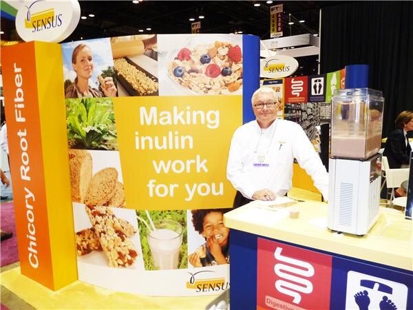 A man with glasses and a white shirt stands at a Sensus booth with a banner reading Making inulin work for you. A Chicory Root Fiber sign and a beverage dispenser are visible.