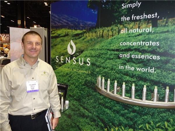 A smiling man in a tan shirt and badge stands in front of a Sensus display showing a green field and the text Simply the freshest, all natural, concentrates and essences in the world.