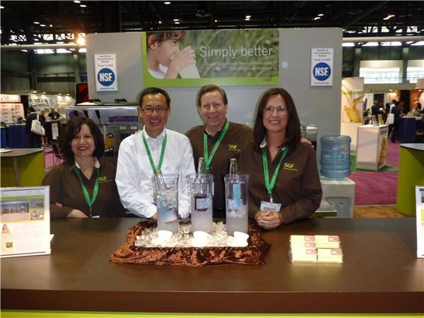 Four people in brown SGF shirts and green lanyards stand behind a counter with glass bottles on ice. Behind them are a Simply better sign and two NSF signs.