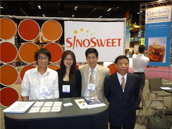 Four people stand behind a counter in front of a SinoSweet sign with orange circles. A CleanSweet banner from JRS International is at right, and printed materials are on the counter.