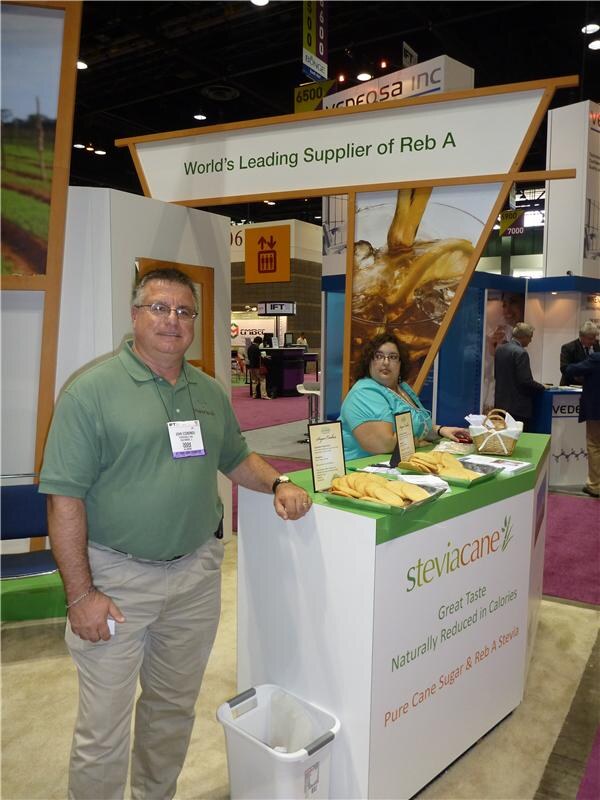 A man in a green polo shirt and badge stands beside a SteviaCane booth counter holding trays of cookies. A seated woman is behind it. A sign above reads World's Leading Supplier of Reb A.