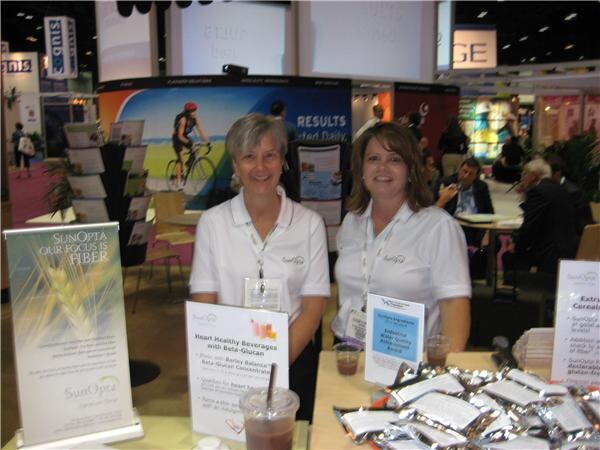 Two smiling women in white SunOpta polo shirts stand at a booth table. Signs mention fiber and heart healthy beverages with beta-glucan, and foil snack packets and cups are on the table.