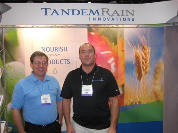 Two men with badges stand in front of a Tandem Rain Innovations booth. One wears a light blue polo and glasses, the other a black polo. Graphics of fruit and wheat appear behind them.