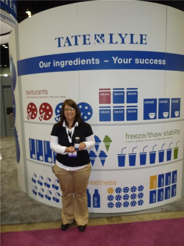 A woman with glasses and a lanyard holds a small bottle in front of a curved white Tate & Lyle display reading 'Our ingredients – Your success', with icons for sauces, cereal and sweetness.