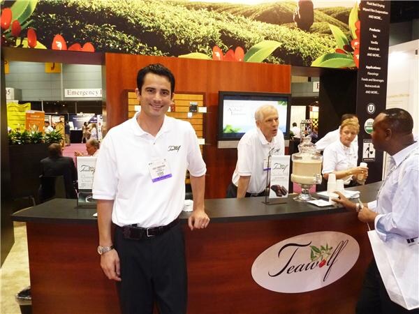 A smiling man in a white polo leans on the Teawolf booth counter. Behind him an older man and a woman stand near a glass tea dispenser, and a visitor in a light blue shirt stands at right.