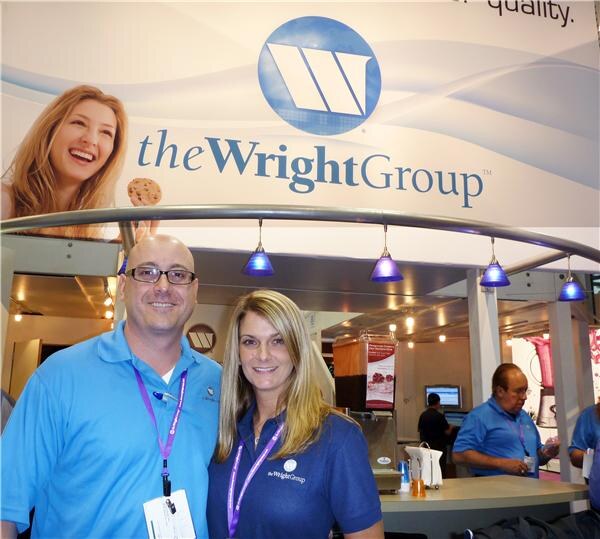 A smiling man with glasses and a woman with long blonde hair, both in blue polo shirts and purple lanyards, pose under a The Wright Group sign. Other staff work at the booth counter behind them.