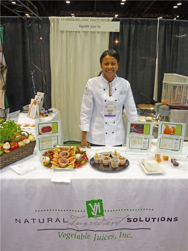 A woman in a white chef's coat stands behind a table with a Vegetable Juices, Inc. banner. The table holds vegetable platters, small sample cups, a basket of produce and product information cards.