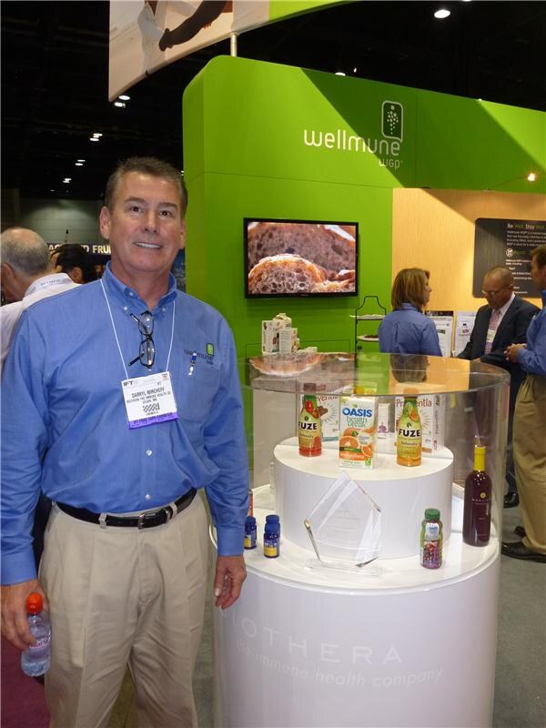 A smiling man in a blue shirt with a name badge stands beside a round display of Fuze and Oasis beverages and supplement bottles, in front of a green Wellmune WGP booth with a television screen.