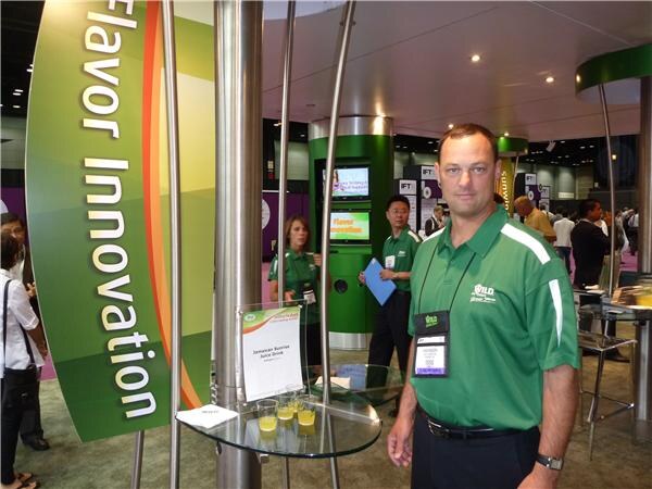A man in a green Wild polo shirt with a name badge stands next to a glass table holding small cups of yellow drink. A 'Flavor Innovation' sign curves at left, and staff stand in the background.