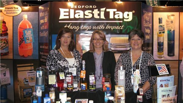 Three women stand behind a table of bottles and jars with hang tags in the Bedford ElastiTag booth, under a sign reading Hang tags with Impact.