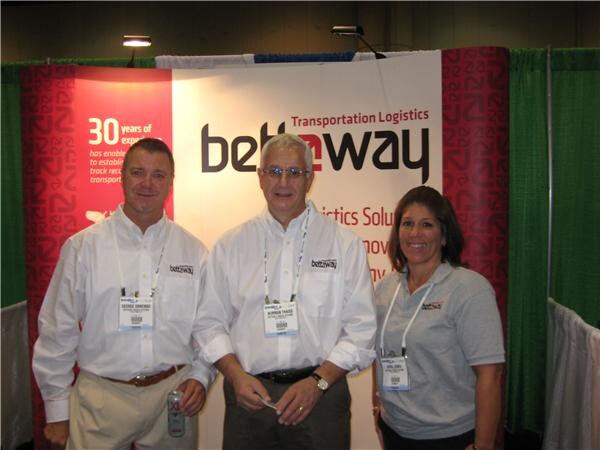 Two men in white shirts and a woman in a gray polo, all wearing lanyards and name badges, stand together in front of a Bettaway Transportation Logistics banner.