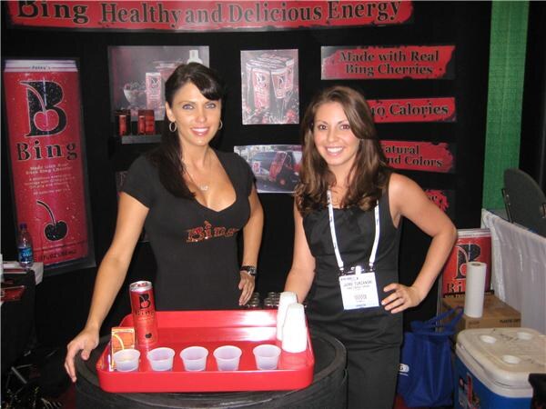 Two smiling women in black tops stand behind a red tray of small sample cups and a Bing can. Behind them are Bing signs reading Healthy and Delicious Energy and Made with Real Bing Cherries.