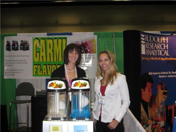 Two women stand behind two granita machines in front of a green Carmi Flavors banner. A Rudolph Research Analytical banner is at the right.