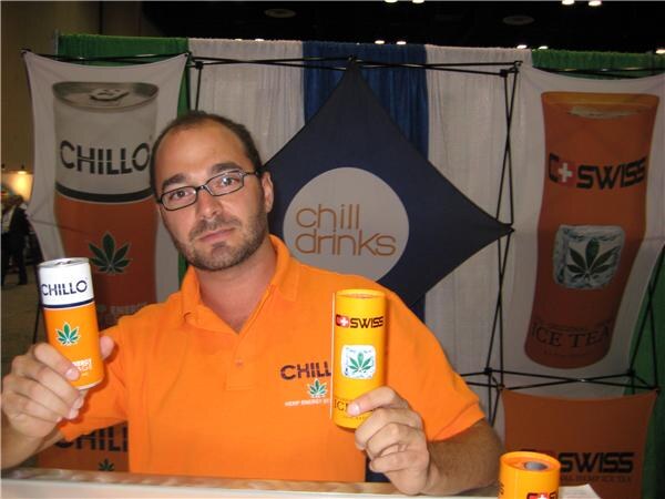A bearded man with glasses in an orange polo holds a Chillo can and a C Swiss can. Behind him are Chill Drinks, Chillo and C Swiss banners.