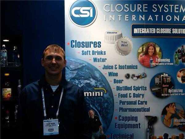 Clint Rush, wearing a dark jacket and a show badge, smiles in front of a Closure Systems International (CSI) display listing closures and capping equipment.