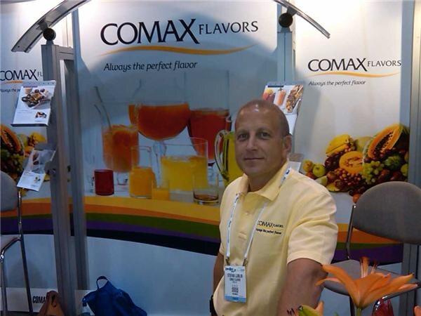 A man in a yellow Comax Flavors polo sits in front of Comax Flavors signs, with glasses of juice and displays of tropical fruit behind him.