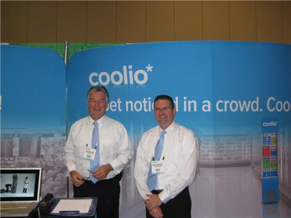 Two men in white shirts and light blue ties stand in front of a blue coolio banner. A laptop and clipboard are on the table in front of them.